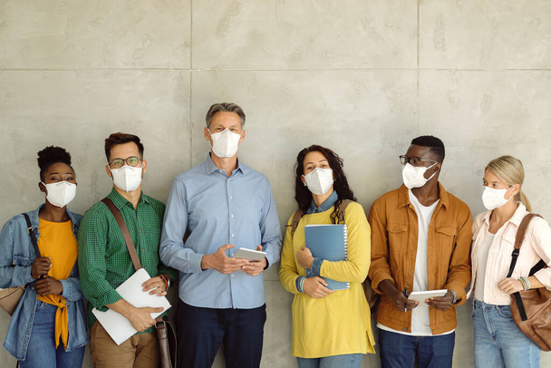 Group of university students and their professor wearing protective face masks while standing against the wall and looking at camera. Copy space.  - Fotoğraf, Görsel