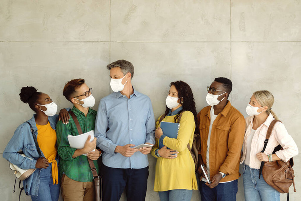 University professor and his students wearing protective face masks while standing by the wall and communicating. Copy space.  - Foto, Bild