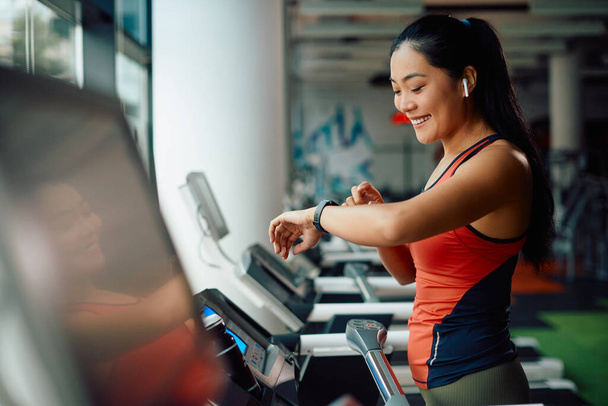 Happy Asian sportvrouw met behulp van slimme horloge tijdens het joggen op loopband tijdens sport training in een sportschool. - Foto, afbeelding