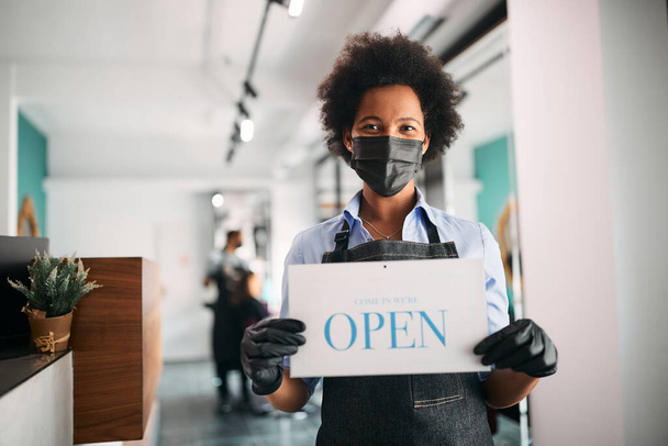 Happy African American hairstylist holding open sign at her salon and wearing face mask due to COVID-19 pandemic. - Photo, image