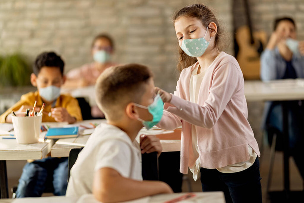 Happy schoolgirl helping her classmate to properly put on a face mask while being at school during coronavirus epidemic.  - 写真・画像