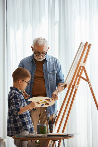 Senior man teaching his grandson to paint on canvas at home studio.  - Foto, Bild