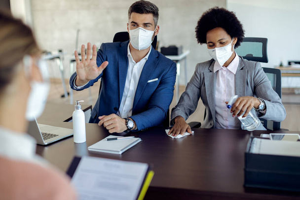 Members of human resource team wearing protective face masks and disinfecting the table before a job interview with potential candidate in the office.  - Фото, изображение