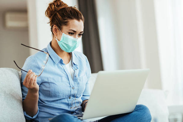 Smiling woman wearing face mask while surfing the net on laptop and relaxing at home during virus epidemic.  - Фото, изображение