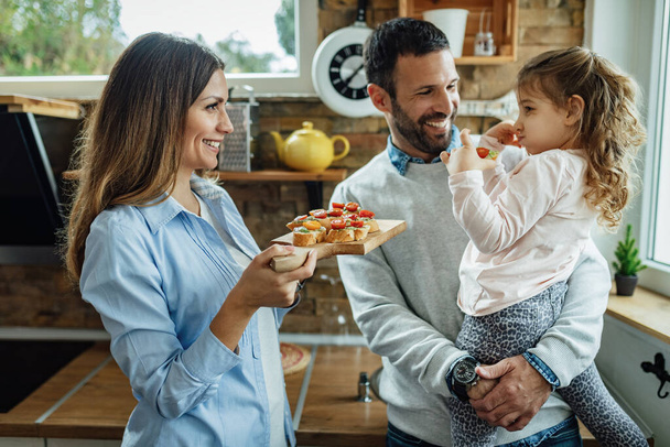 Mladí šťastní rodiče a jejich malá dcera komunikovat při jídle bruschetta v kuchyni.  - Fotografie, Obrázek
