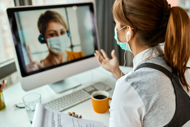 Zakenvrouw met gezichtsmasker communiceert met haar collega via videogesprek op kantoor tijdens COVID-19 pandemie.  - Foto, afbeelding