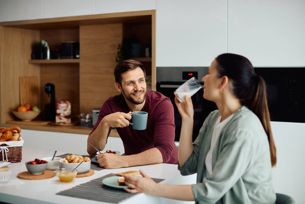 Young happy man dinking coffee and communicating with his wife while having breakfast together at home. - Foto, Bild