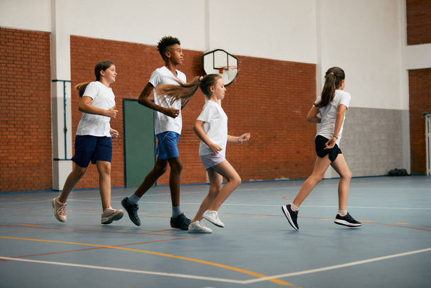 Full length of elementary students running while having PE class at school gym. - Foto, afbeelding
