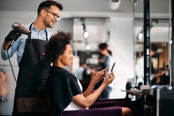 Щаслива афро-американська жінка, що показує щось на смартфоні своєму перукарю під час стилізації волосся в салоні.  - Фото, зображення