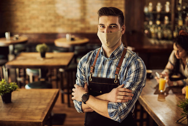 Portrait of waiter standing with arms crossed and wearing protective face mask in a pub during coronavirus pandemic.  - Foto, immagini