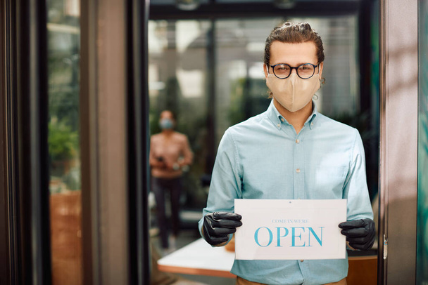 Happy waiter wearing protective face mask and holding open sign at reopened restaurant during COVID-19 epidemic - 写真・画像