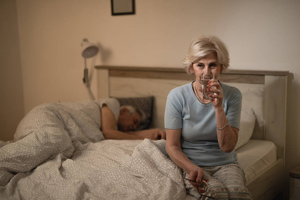 Senior woman having a glass of water in bedroom at night while her husband is sleeping.  - Zdjęcie, obraz