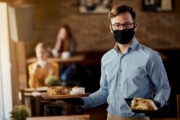 Молодой счастливый официант носит защитную маску для лица, подавая еду в ресторане.  - Фото, изображение