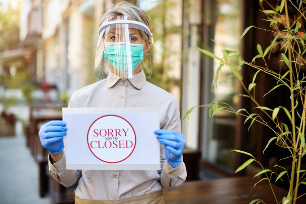 Cafe owner holding closed sign while wearing protective face mask and visor due to coronavirus epidemic.  - Foto, Bild
