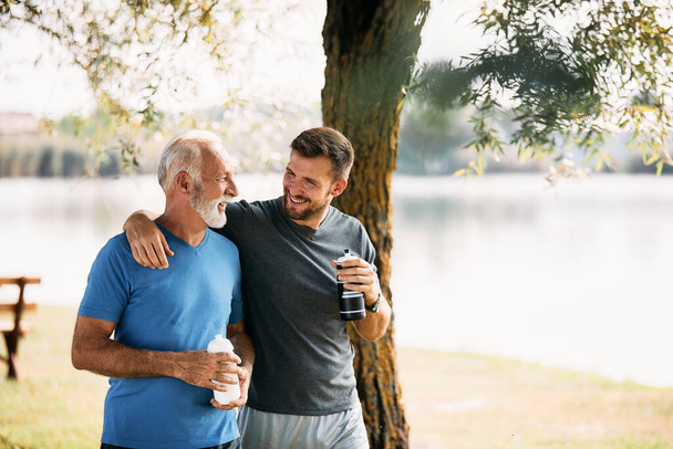 Happy athletic man and his mature father talking while drinking water and walking after sports training in nature. Copy space. - Fotografie, Obrázek