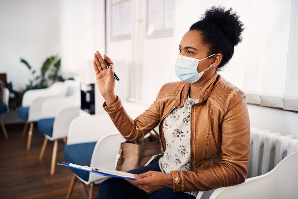 African American woman raising hand to ask something while writing her data in dental record at dental clinic. She is wearing face mask due to coronavirus pandemic. - Zdjęcie, obraz