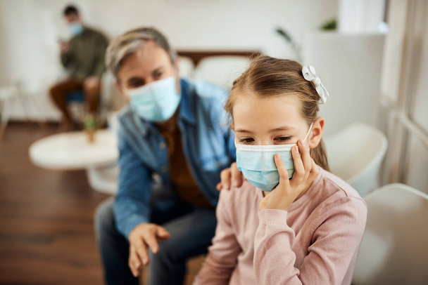 Little girl feeling nervous before dental exam while sitting with her father in waiting room and wearing protective face mask due to COVID-19 pandemic.  - Foto, Imagem