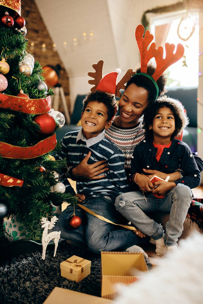 Happy black mother embracing her children while sitting by Christmas tree and enjoying in holidays at home.  - 写真・画像
