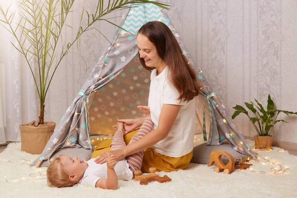 Indoor shot of young mother playing with with baby daughter on floor near kids wigwam, expressing positive emotions, spending time with cute kid in peetee tent at home. - Фото, зображення