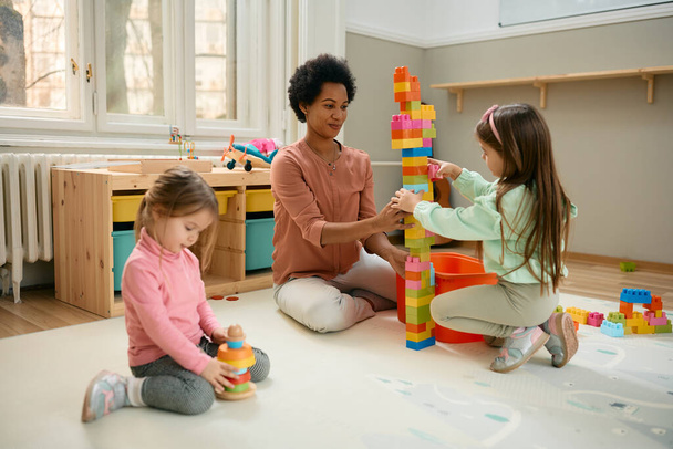 African American teacher and little girls stacking toy blocks while playing on the floor at kindergarten.  - Фото, изображение