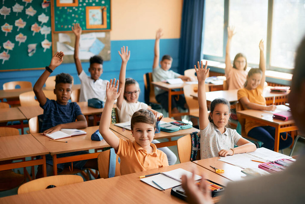 Μεγάλη ομάδα από χαρούμενα παιδιά που σηκώνουν τα χέρια για να απαντήσουν στην ερώτηση του δασκάλου για μια τάξη στην τάξη.  - Φωτογραφία, εικόνα