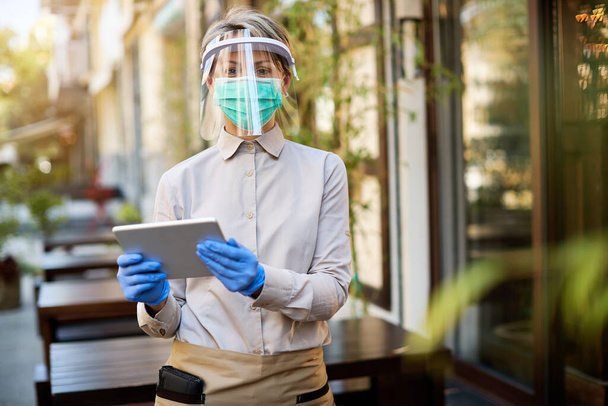 Ευτυχισμένη σερβιτόρα που χρησιμοποιεί digital tablet ενώ φοράει visor και προστατευτική μάσκα προσώπου σε υπαίθριο καφέ,  - Φωτογραφία, εικόνα