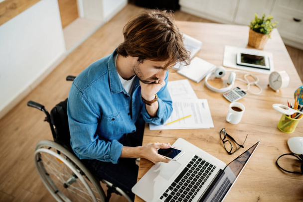 Високий кут зору молодого підприємця в інвалідному візку на смартфон під час роботи на комп'ютері вдома
.  - Фото, зображення