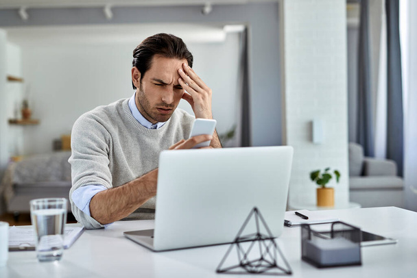 Distraught entrepreneur working at home and holding his head in pain while reading text message on cell phone.  - Foto, Imagem