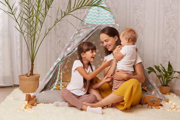 Indoor shot of Caucasian attractive woman with her children sitting together teepee tent, mom playing with daughters, spending weekend with kids while playing in wigwam. - Fotografie, Obrázek