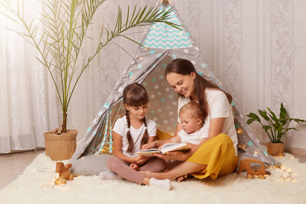 Caring mother and her daughters sitting near wigwam with book in hands, woman and kids playing at home, reading fairy tales and interesting stories, spending time together. - 写真・画像