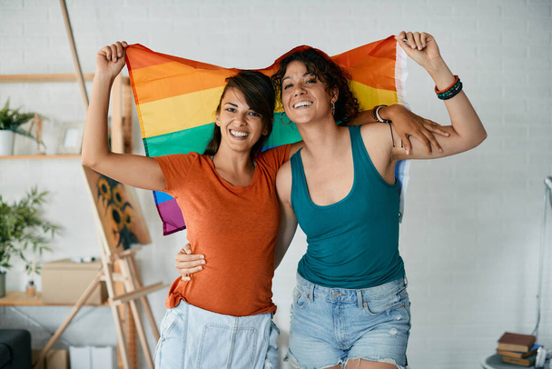 Young happy lesbian couple having fun and holding rainbow flag at home while looking at camera. - Φωτογραφία, εικόνα