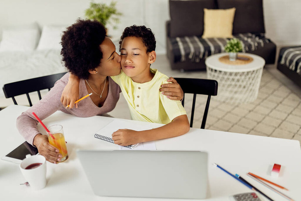 Caring black mother kissing her small boy while after finishing homework in the living room.  - Foto, Bild