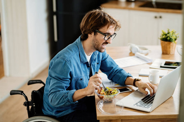 Молодий щасливий підприємець у інвалідному кріслі, що серфінгує мережу на ноутбуці, в той час як здорова їжа вдома
.  - Фото, зображення