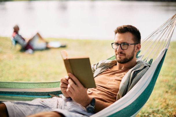 Uvolněný muž sedí v houpací síti a čte knihu během kempování v přírodě. - Fotografie, Obrázek