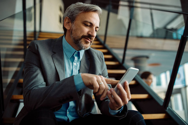 Επιχειρηματίας που χρησιμοποιεί το κινητό τηλέφωνο και γραπτών μηνυμάτων, ενώ κάθεται σε εσωτερική σκάλα.  - Φωτογραφία, εικόνα