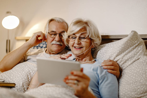 Happy senior woman and her husband using digital tablet while relaxing in bed at home.   - Fotó, kép