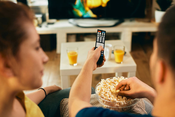 Крупним планом чоловік змінює канали по телевізору і їсть попкорн, розслабляючись зі своєю дівчиною вдома
.  - Фото, зображення
