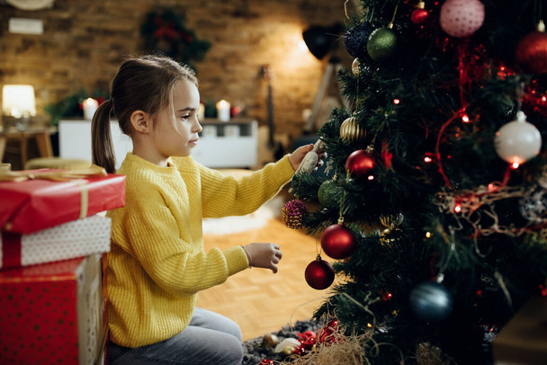 Cute little girl preparing for holidays and decorating Christmas tree at home.  - Fotó, kép