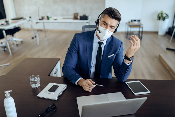 Happy entrepreneur using laptop while having video call in the office. He is wearing protective face mask due to coronavirus epidemic.  - Foto, Imagen