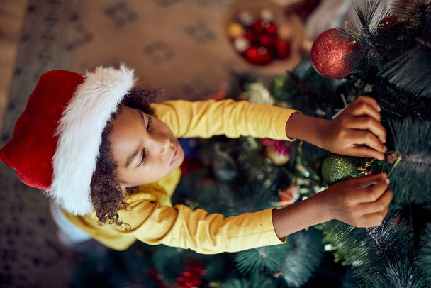 High angle view of small black girl decorating Christmas tree at home.  - Foto, Bild