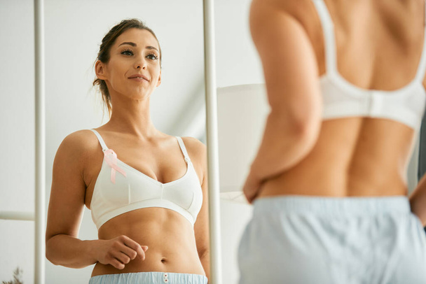 Віддзеркалення у дзеркалі молодої жінки зі стрічкою обізнаності про рак молочної залози у її бюстгальтері вдома
. - Фото, зображення