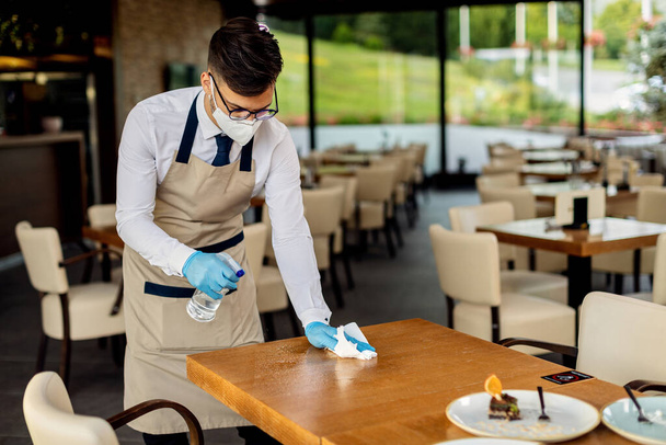 Waiter wearing protective face mask and gloves while disinfecting cafe tables after work during virus epidemic.  - Foto, afbeelding