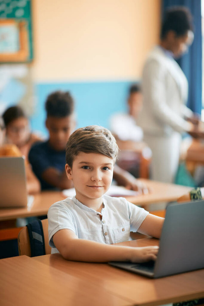 Χαμογελώντας μαθητής δημοτικού χρησιμοποιώντας φορητό υπολογιστή, ενώ e-learning κατά τη διάρκεια του μαθήματος στην τάξη και κοιτάζοντας κάμερα.  - Φωτογραφία, εικόνα