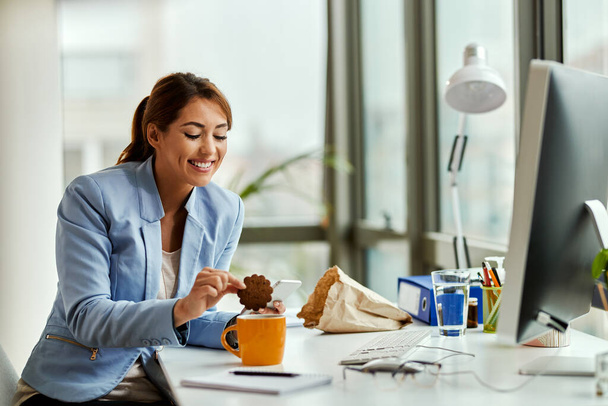 Усміхнена бізнес-леді занурює печиво в чашку молока під час роботи в офісі
.  - Фото, зображення