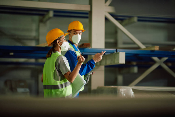 Two warehouse workers communicating while walking through storage room and wearing protective face masks.  - Photo, Image
