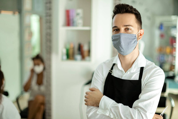 Young hairdresser with protective face mask standing with arms crossed at hair salon after reopening during coronavirus epidemic.  - Foto, Bild