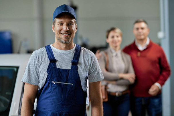Portret van een gelukkige automonteur in de garage. Zijn klanten staan op de achtergrond.  - Foto, afbeelding