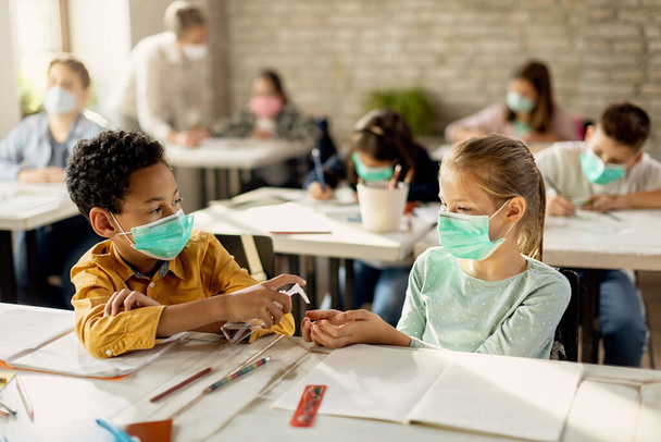 African American schoolboy spraying classmate's hands with disinfectant while wearing protective face masks on a class in the classroom.  - 写真・画像