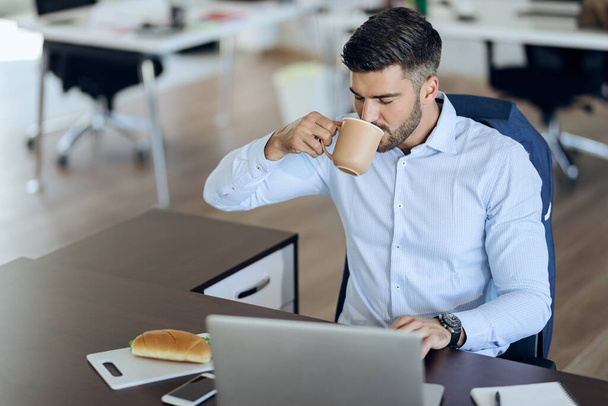 Молодий підприємець насолоджується чашкою кави під час роботи на ноутбуці в офісі
. - Фото, зображення