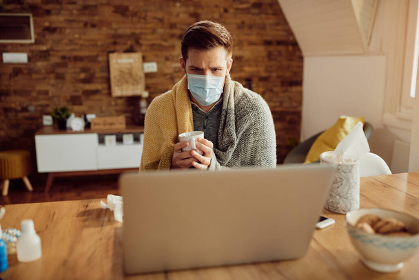 Больной человек, завернутый в одеяло, пьет чай, пользуясь компьютером и надевая маску для лица дома. - Фото, изображение